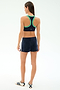 A woman faces away in a dark sports bra with green trim, Heritage Velour Short - Indigo/White by SPLITS59, and white Adidas sneakers, all shown on a plain white background.