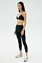 A woman stands against a plain background wearing the SPLITS59 Theo Rigor Bra - Black, paired with black leggings and silver athletic shoes—ideal for versatile sport performance.