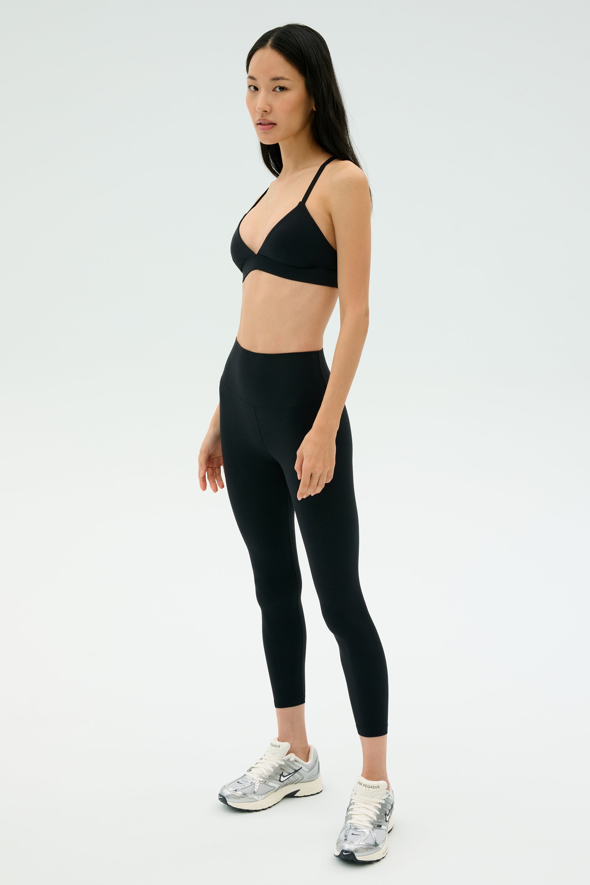 A woman stands against a plain background wearing the SPLITS59 Theo Rigor Bra - Black, paired with black leggings and silver athletic shoes—ideal for versatile sport performance.