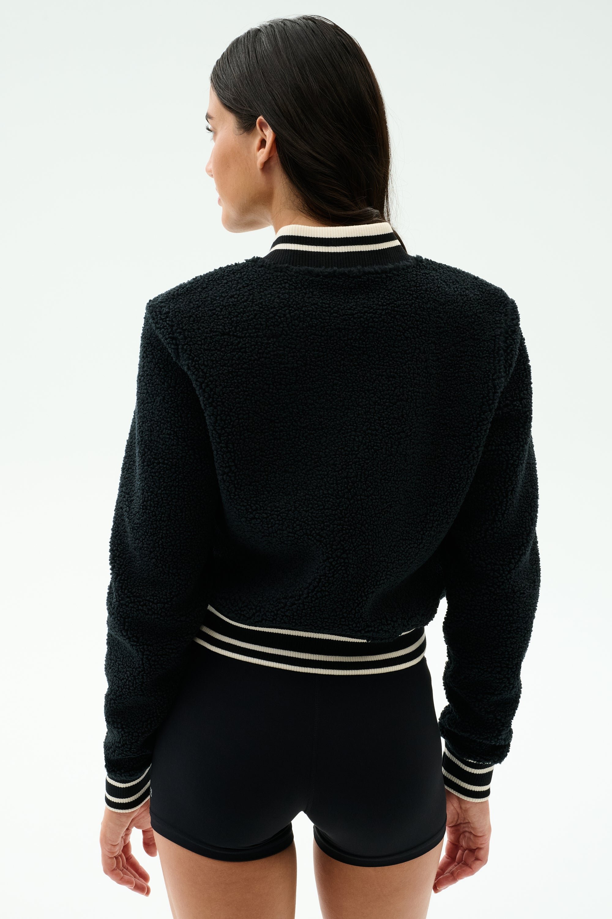 A person faces away wearing the SPLITS59 Willa Sherpa Jacket in black, featuring a textured baseball style with white stripes on the collar, cuffs, and waistband, paired with black shorts against a plain light background.