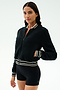 A woman stands against a plain background wearing the SPLITS59 Willa Sherpa Jacket in Black with striped cuffs and black fitted shorts—an ideal combo for winter workouts.