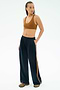 A woman stands with one hand in her pocket, wearing the SPLITS59 Airweight Scoop Back Bra in Honey, navy blue track pants with a brown stripe, and athletic shoes, against a white background.
