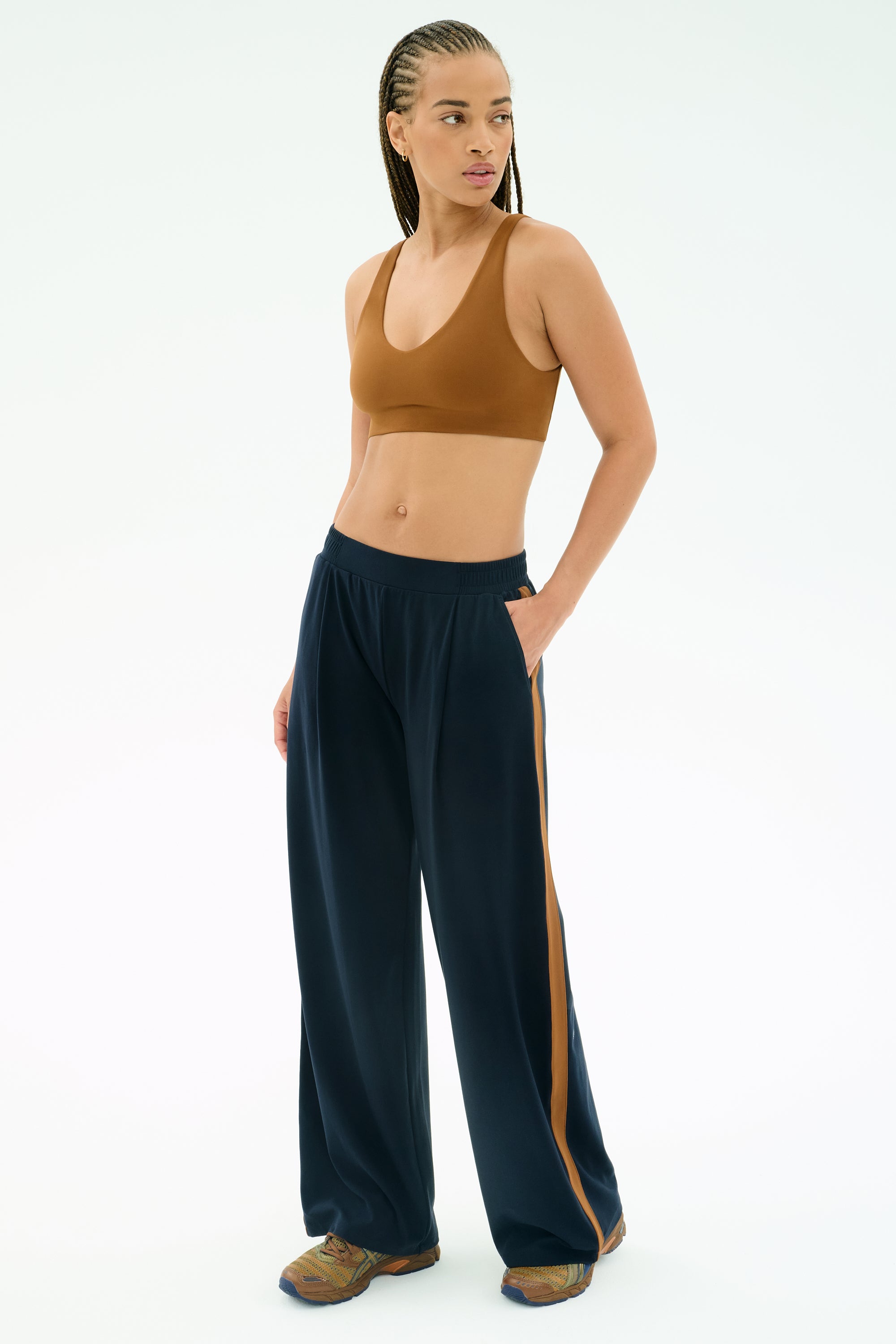 A woman stands with one hand in her pocket, wearing the SPLITS59 Airweight Scoop Back Bra in Honey, navy blue track pants with a brown stripe, and athletic shoes, against a white background.
