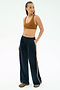 A woman stands with one hand in her pocket, wearing the SPLITS59 Airweight Scoop Back Bra in Honey, navy blue track pants with a brown stripe, and athletic shoes, against a white background.