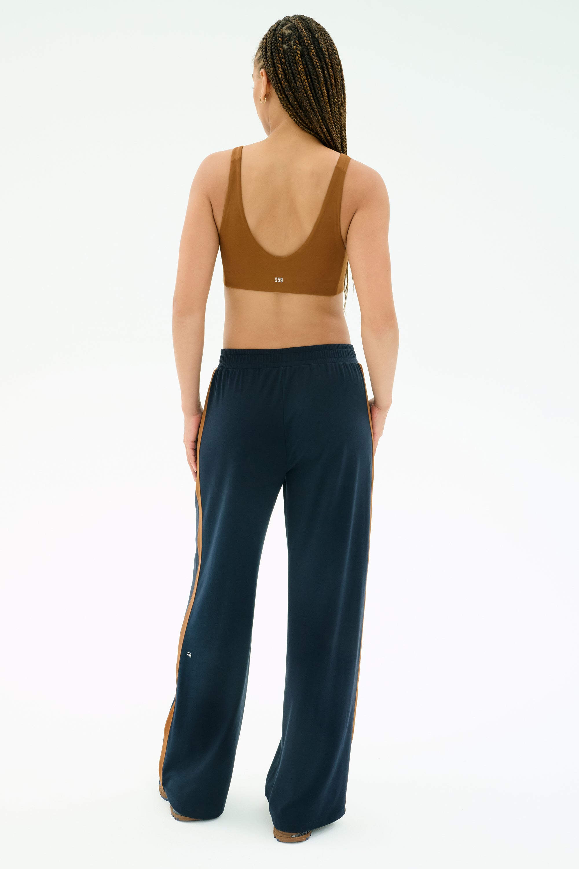 A person with braided hair stands facing away, wearing the SPLITS59 Airweight Scoop Back Bra in Honey and navy blue track pants with a stripe down the side.