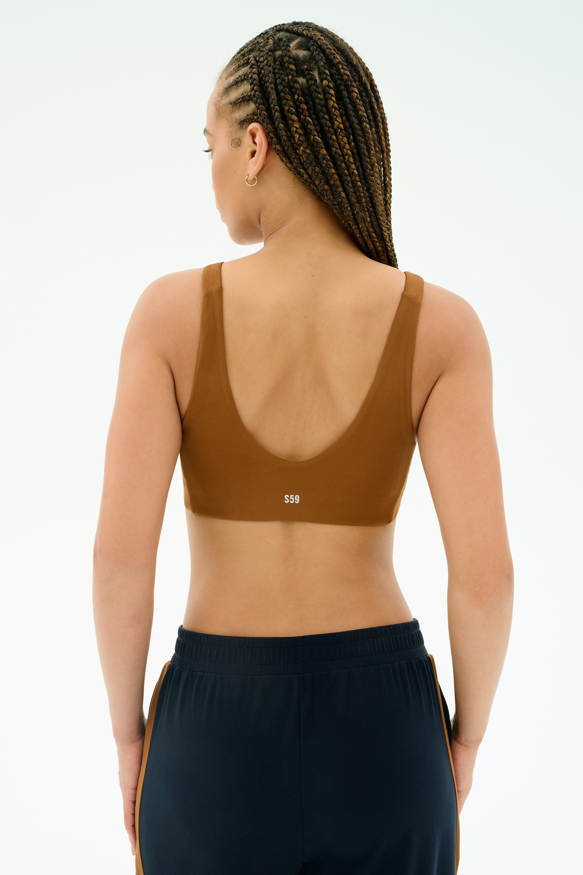 A person with braided hair is shown from the back wearing the SPLITS59 Airweight Scoop Back Bra in Honey and dark athletic pants against a plain background.