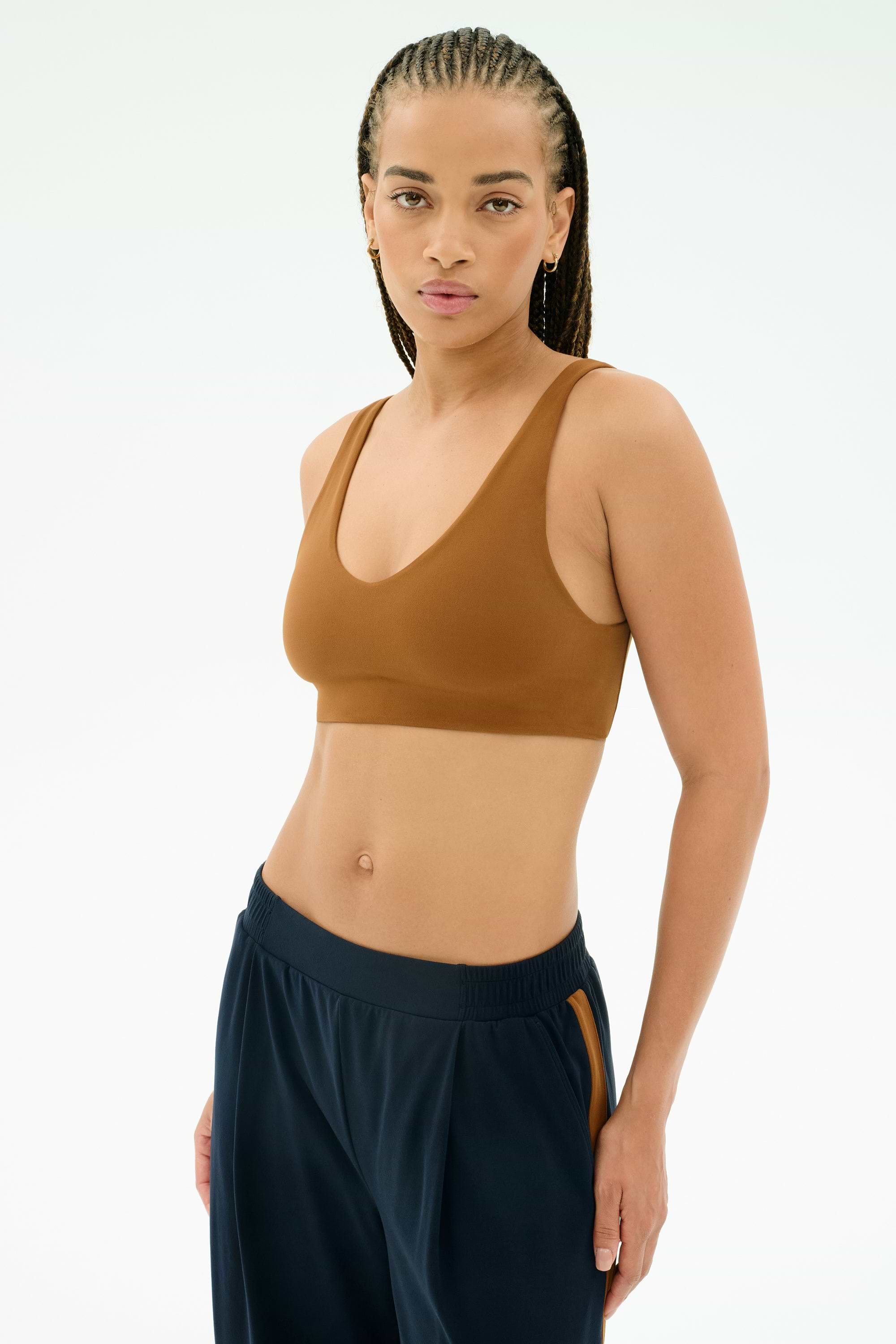 A person wearing the SPLITS59 Airweight Scoop Back Bra in Honey and dark blue jogger pants stands against a plain light background, looking directly at the camera.