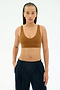 A woman with long braids wears the SPLITS59 Airweight Scoop Back Bra in Honey and dark pants, standing against a plain white background with her hands in her pockets.