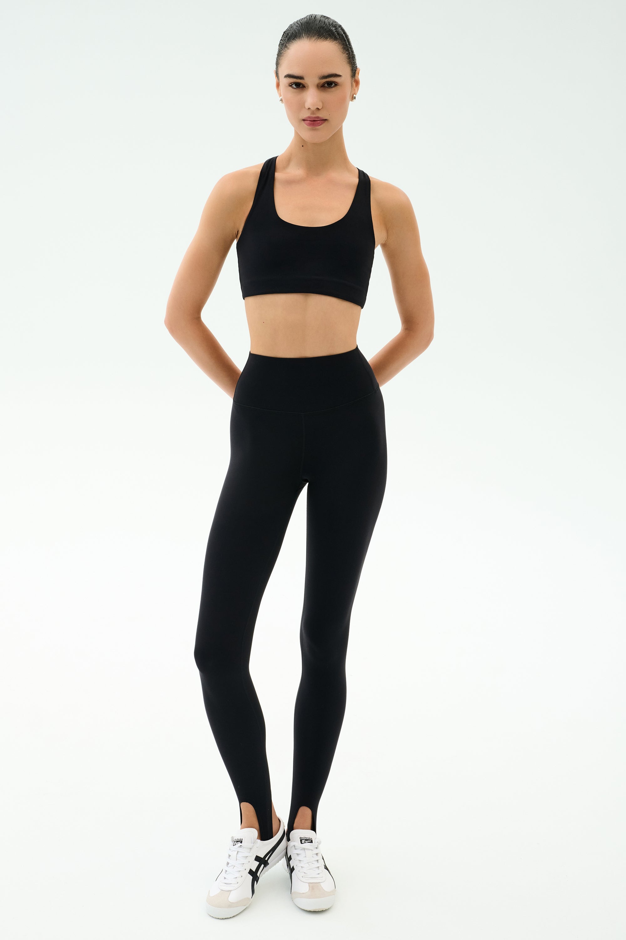 A person stands facing forward in a SPLITS59 Sara Airweight Bra - Black, black high-waisted leggings, and white sneakers—an ideal Pilates or hot yoga look—against a plain white background.