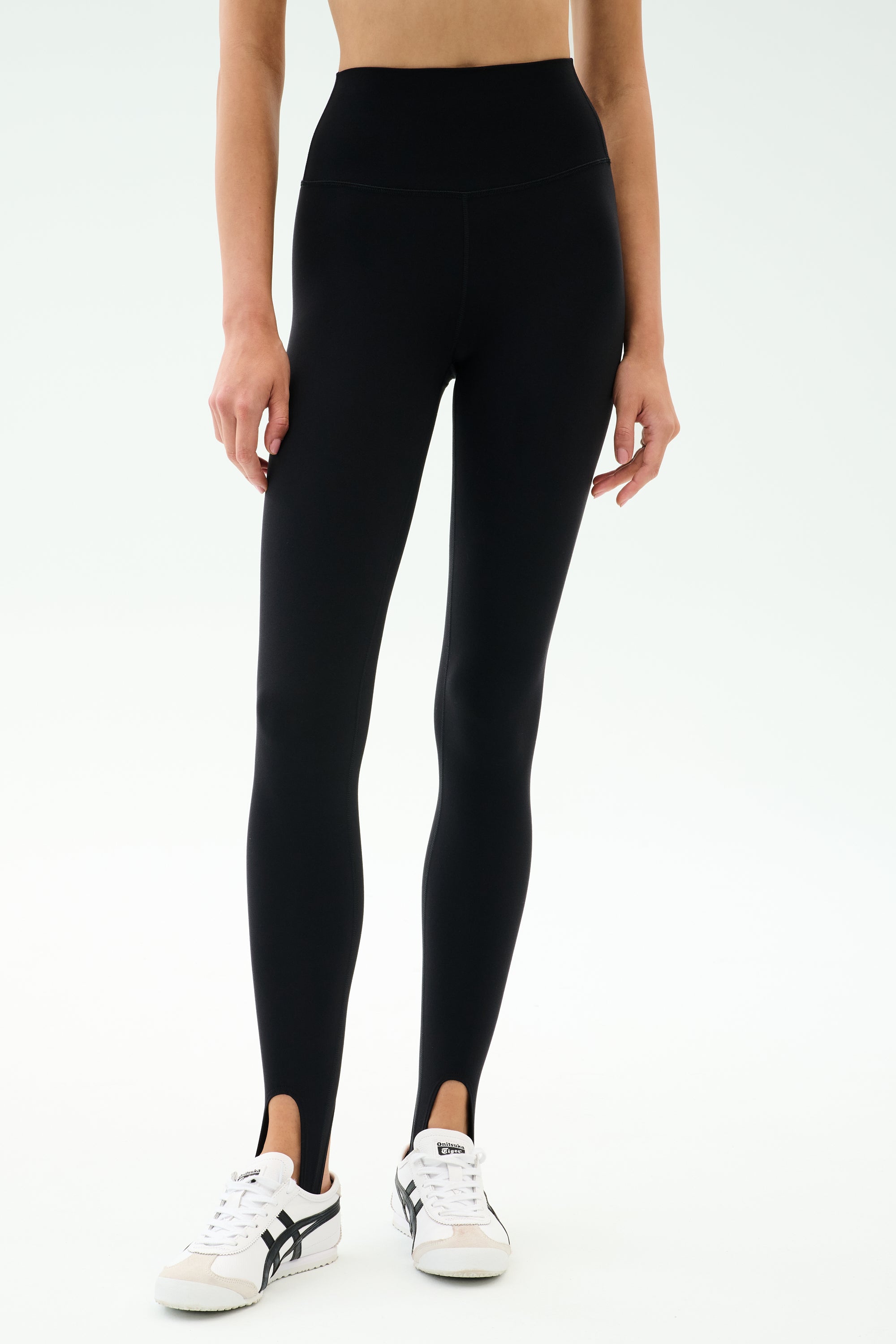 A person wearing SPLITS59 River High Waist Airweight Stirrup leggings in black with white sneakers, standing against a plain light background.