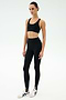 A person stands against a plain background wearing the SPLITS59 Sara Airweight Bra in black, paired with black high-waisted stirrup leggings—ideal for Pilates or hot yoga—and white athletic sneakers.