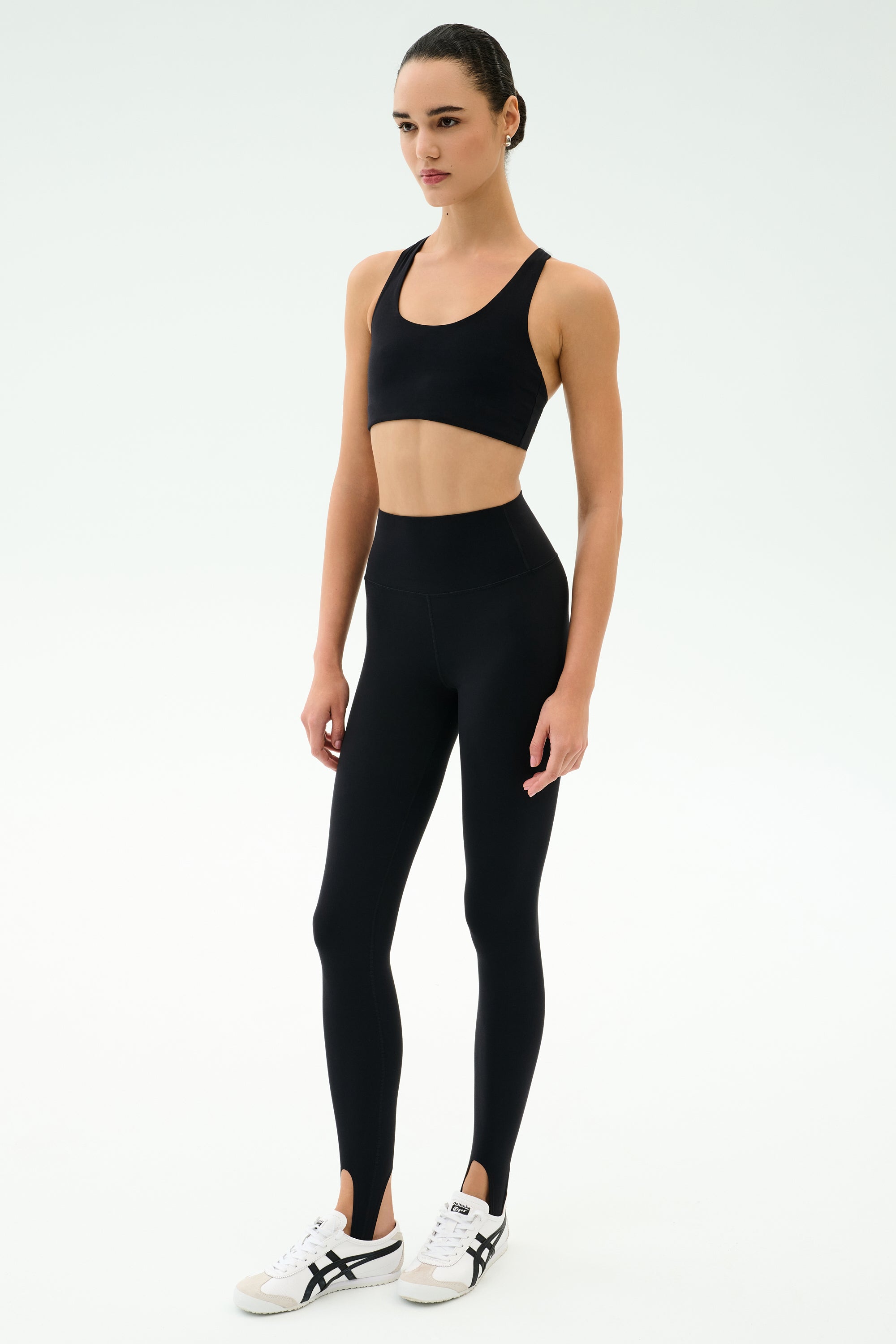 A person stands against a plain background wearing the SPLITS59 Sara Airweight Bra in black, paired with black high-waisted stirrup leggings—ideal for Pilates or hot yoga—and white athletic sneakers.