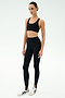 A person stands against a plain background wearing the SPLITS59 Sara Airweight Bra in black, paired with black high-waisted stirrup leggings—ideal for Pilates or hot yoga—and white athletic sneakers.