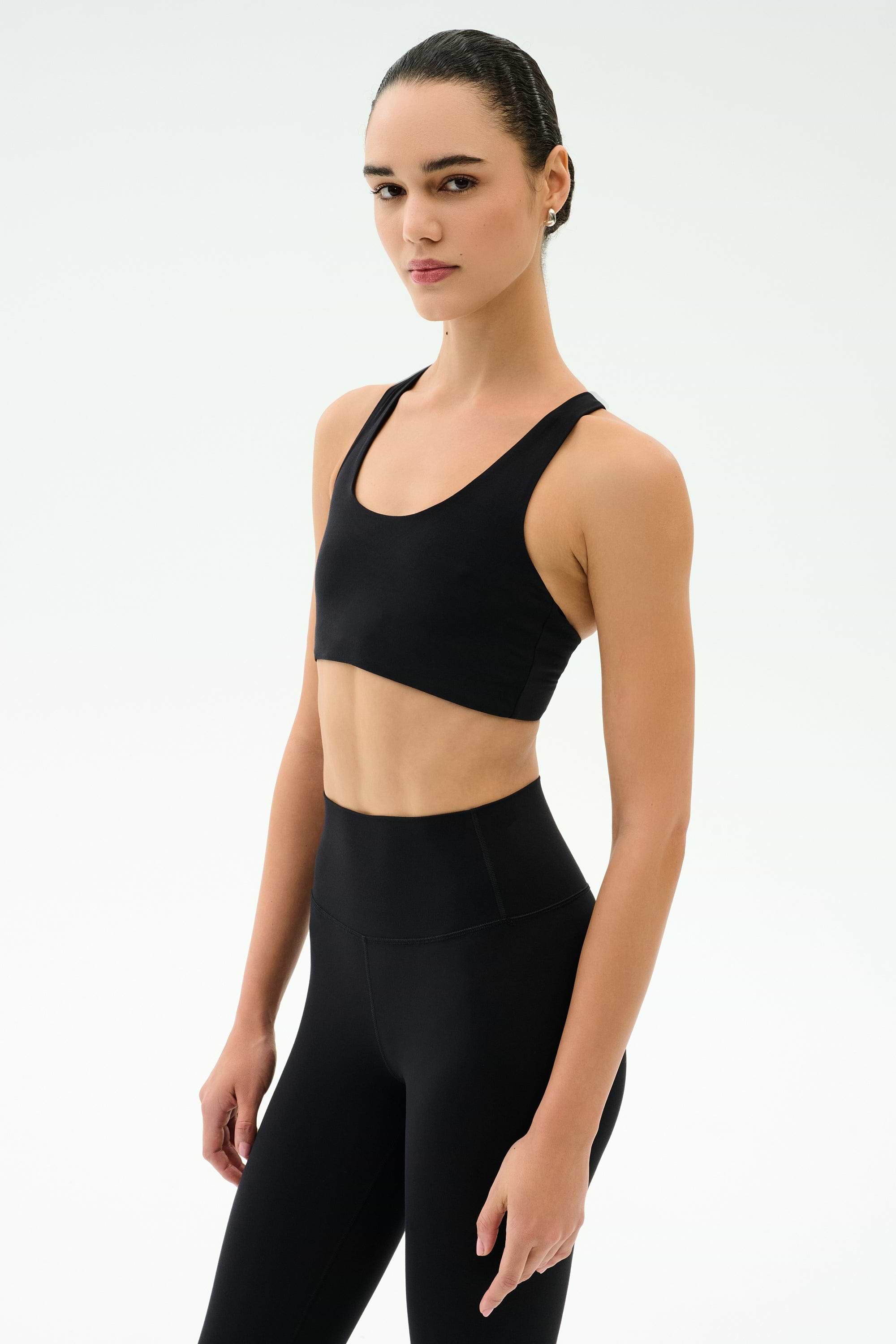 A woman in the SPLITS59 Sara Airweight Bra - Black and high-waisted leggings stands against a plain white backdrop, gazing at the camera—ideal activewear for Pilates or hot yoga.
