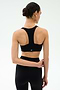 A woman with dark hair in a bun wears the SPLITS59 Sara Airweight Bra in black and matching leggings, standing with her back to the camera against a light background—ideal for Pilates or hot yoga.