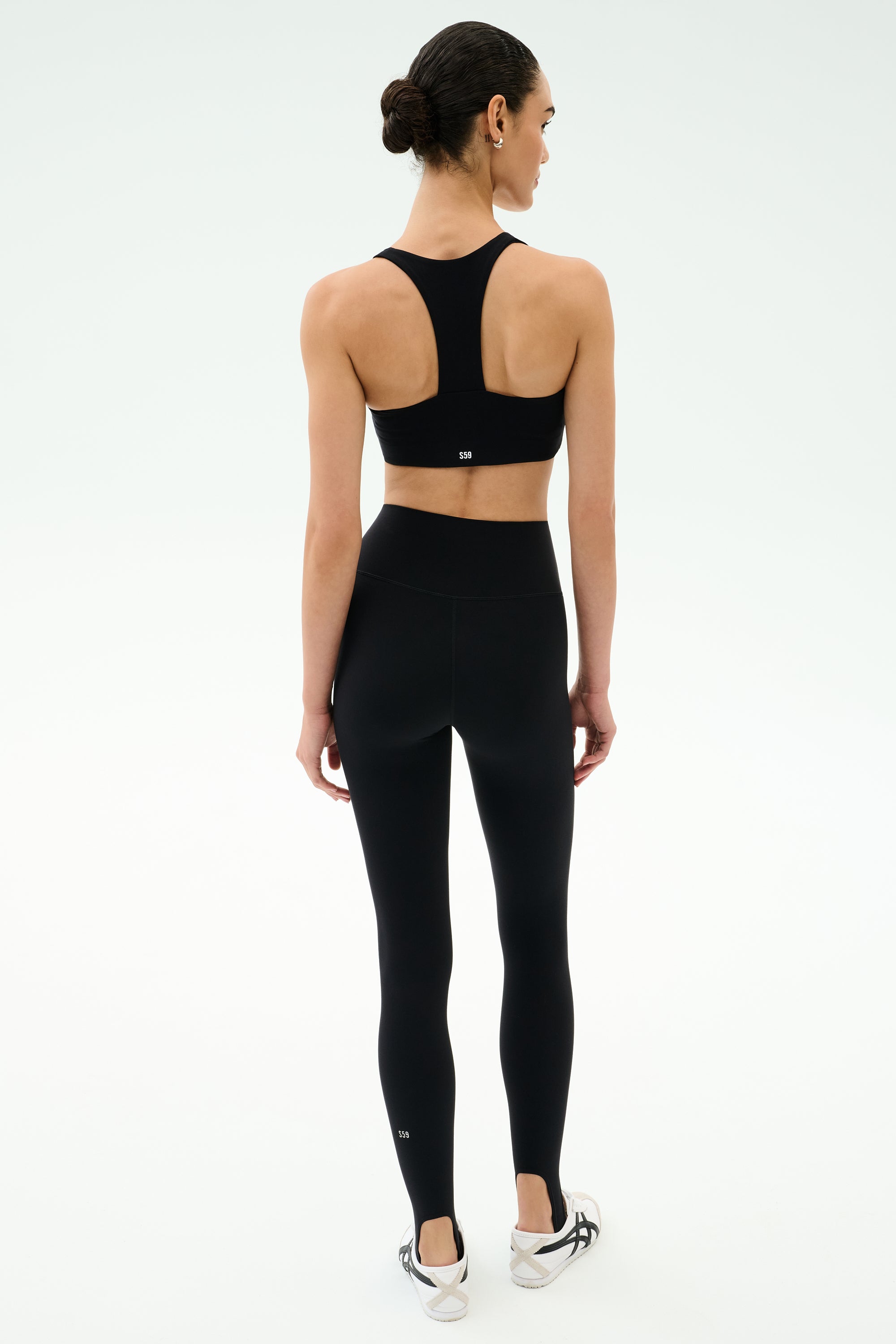 A woman, seen from behind, wears the SPLITS59 Sara Airweight Bra in black with matching leggings and athletic shoes—ideal for Pilates or hot yoga—set against a plain white background.