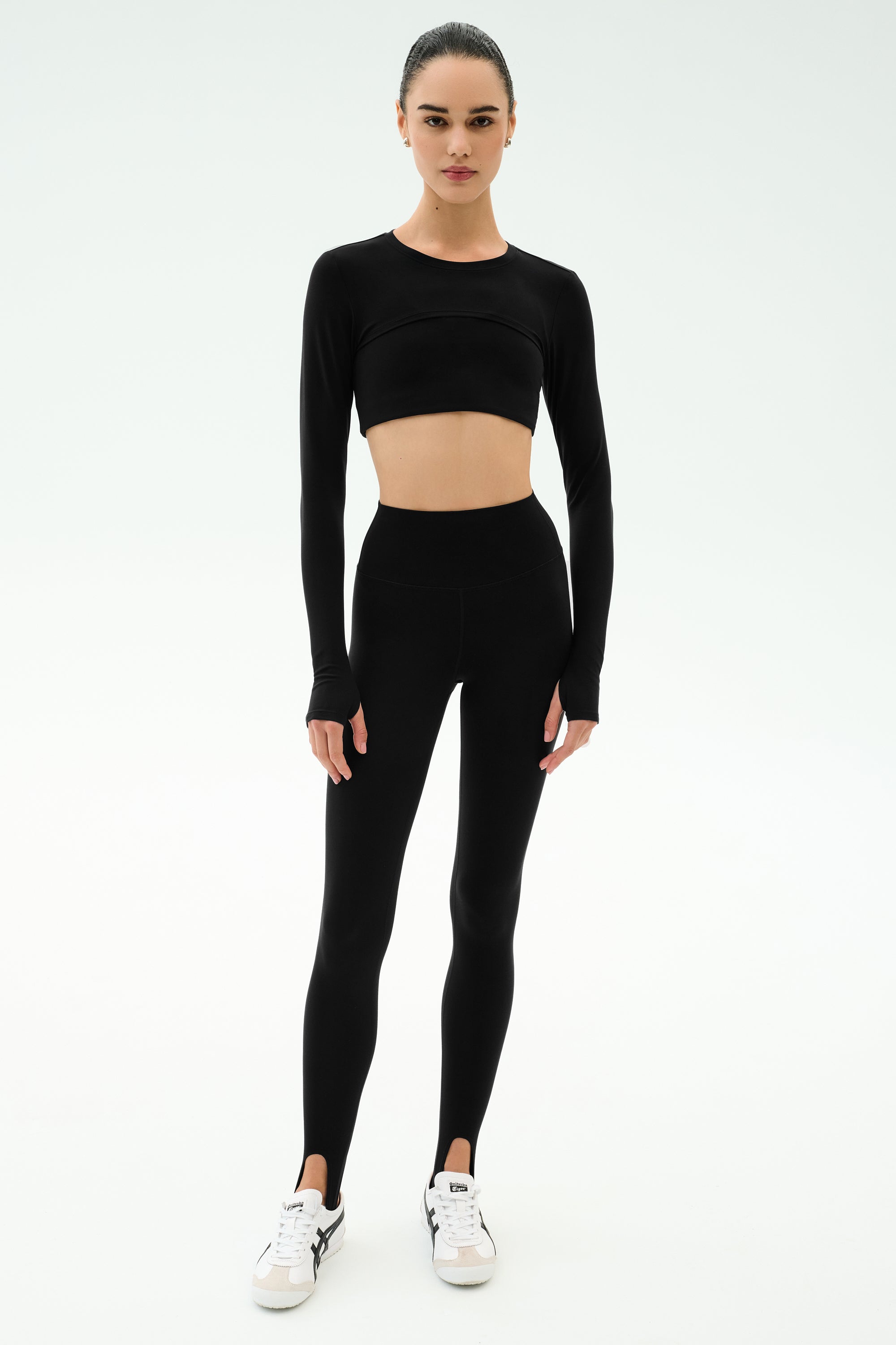 A person stands facing forward, wearing the SPLITS59 Amelia Airweight Lite Shrug in black over a long-sleeve crop top, black high-waisted stirrup leggings, and white sneakers against a plain white background.