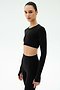 A woman stands against a plain background wearing the SPLITS59 Amelia Airweight Lite Shrug in black, layered over a matching long-sleeve crop top and high-waisted leggings—ideal for yoga or dance warmup sessions.