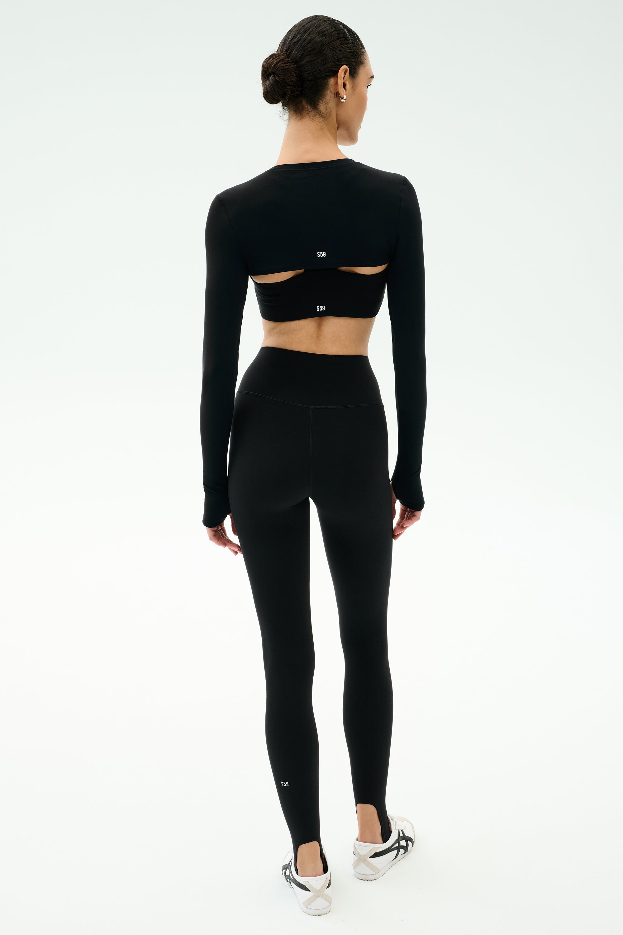 A person stands facing away, wearing the SPLITS59 Amelia Airweight Lite Shrug in black with matching high-waisted leggings, short hair, and white sneakers.