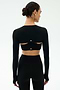 A woman in a fitted black long-sleeve crop top with open back cutouts and matching high-waisted leggings, styled with the SPLITS59 Amelia Airweight Lite Shrug in Black, stands against a plain light background.