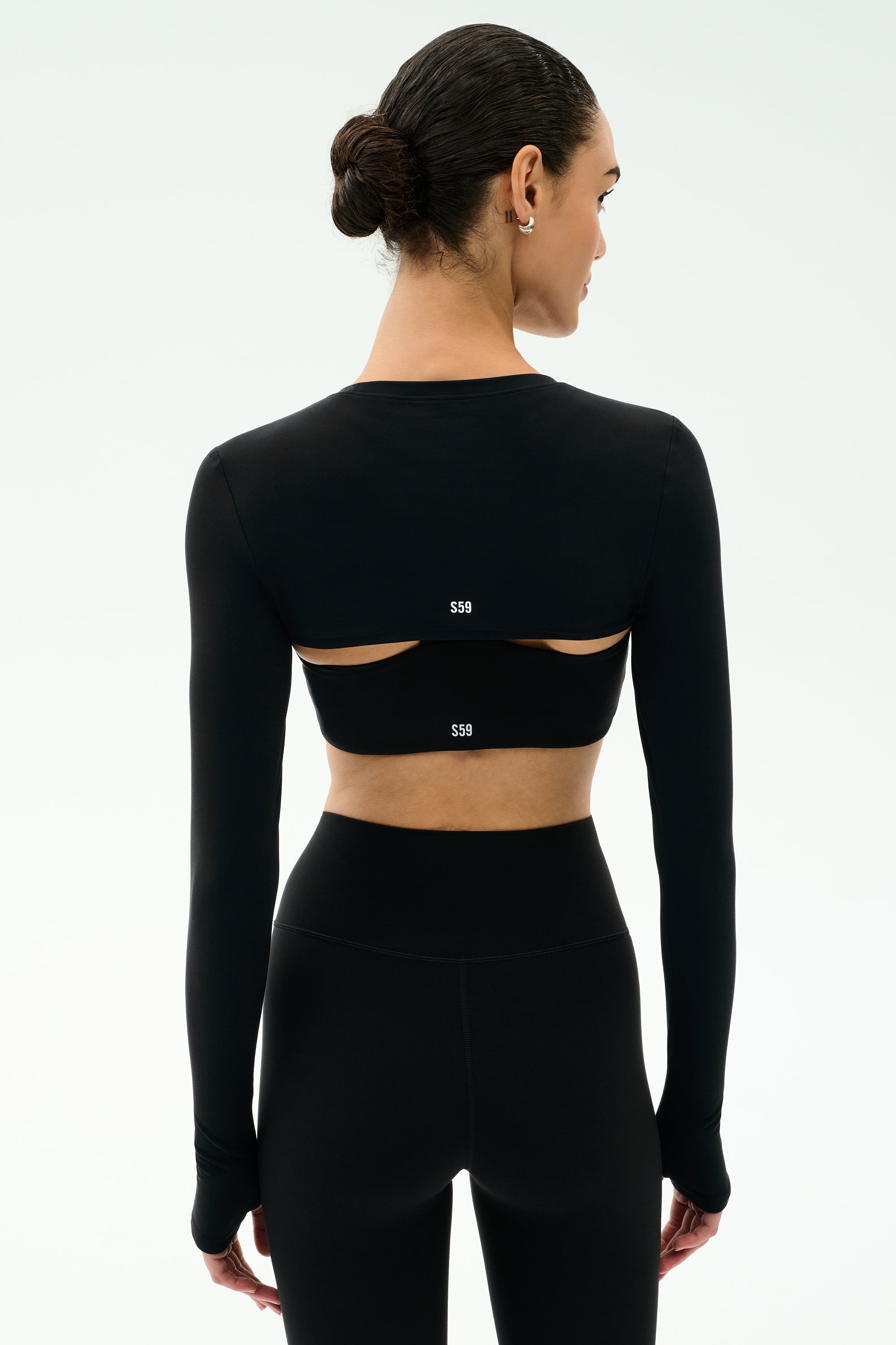 A woman in a fitted black long-sleeve crop top with open back cutouts and matching high-waisted leggings, styled with the SPLITS59 Amelia Airweight Lite Shrug in Black, stands against a plain light background.