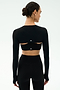A woman in a fitted black long-sleeve crop top with open back cutouts and matching high-waisted leggings, styled with the SPLITS59 Amelia Airweight Lite Shrug in Black, stands against a plain light background.