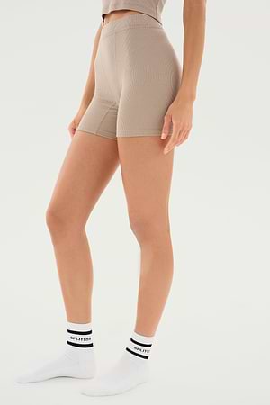 A person wearing Theo Rib Short - Latte from SPLITS59 in beige high-waisted style and white ankle socks with black writing stands against a white background, showcasing a classic design that exudes tailored comfort.