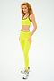 A person with curly hair, styled in a bright yellow workout outfit and sneakers, stands ready for gym workouts against a plain background. They're wearing the Easton Rigor High Waist 7/8 - Chartreuse/Black by SPLITS59.
