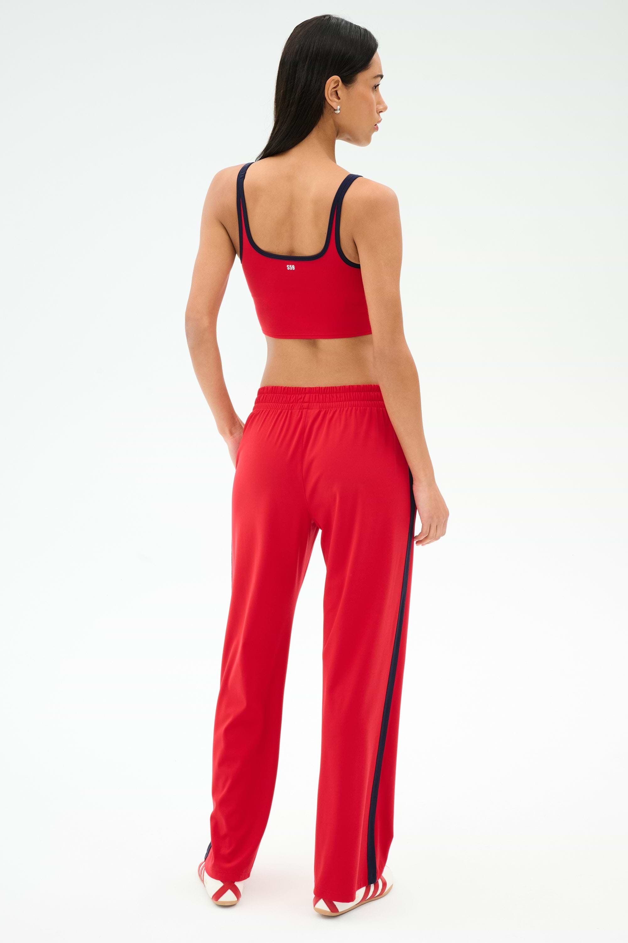 A woman faces away, wearing a red sports bra and SPLITS59 Maxie Rigor Track Pant in Pirate Red/Indigo—wide leg track pants with indigo trim—against a plain light background.