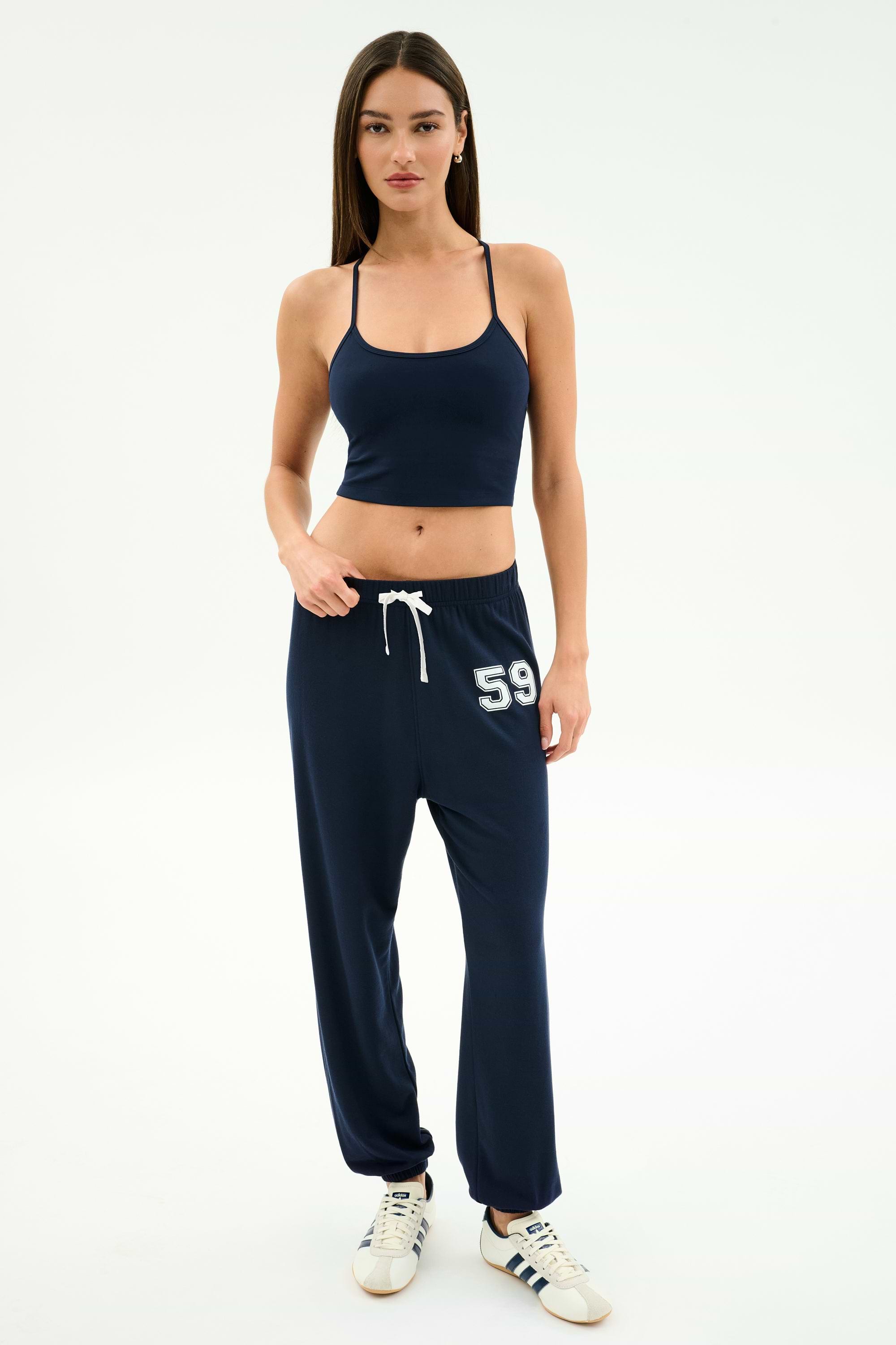 A woman stands against a plain background wearing the SPLITS59 Airweight Crop - Indigo with matching navy joggers featuring "59" on the thigh and white sneakers—a stylish look for hot yoga or casual outings.