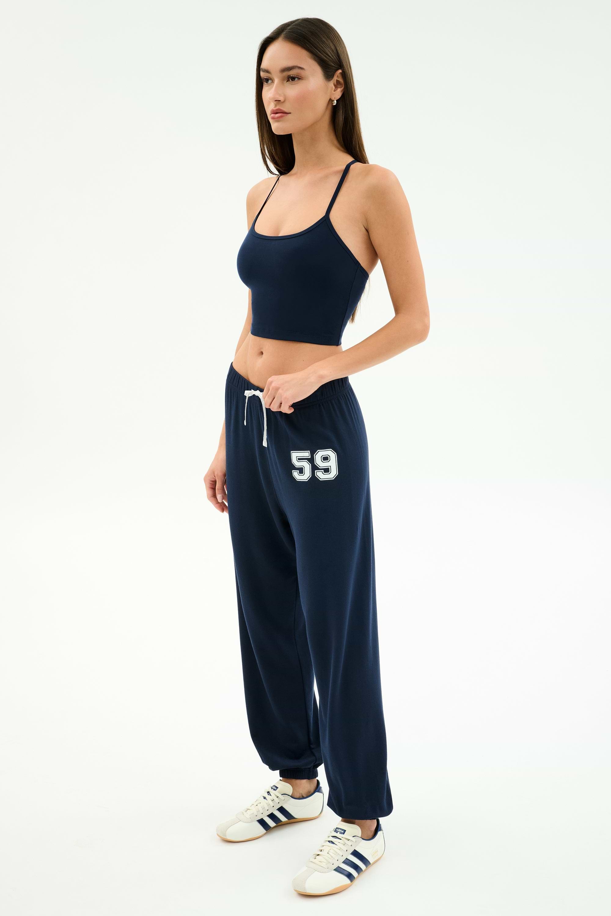 A woman wears the SPLITS59 Airweight Crop - Indigo with matching drawstring sweatpants featuring “59” on the left leg, plus white sneakers with navy stripes—ideal for hot yoga or casual outings.
