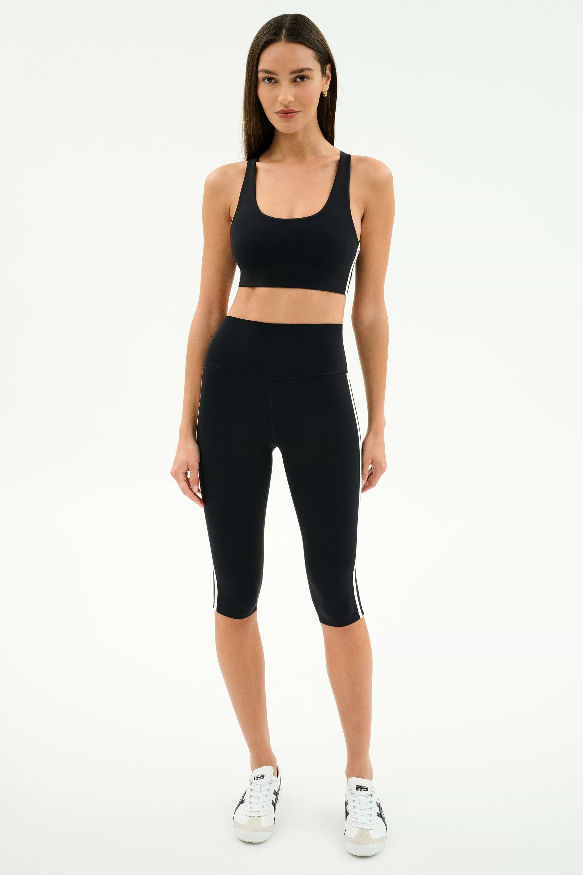 A woman stands against a plain background wearing the SPLITS59 Ella High Waist Airweight Capri in Black/White, a black sports bra, and white sneakers.