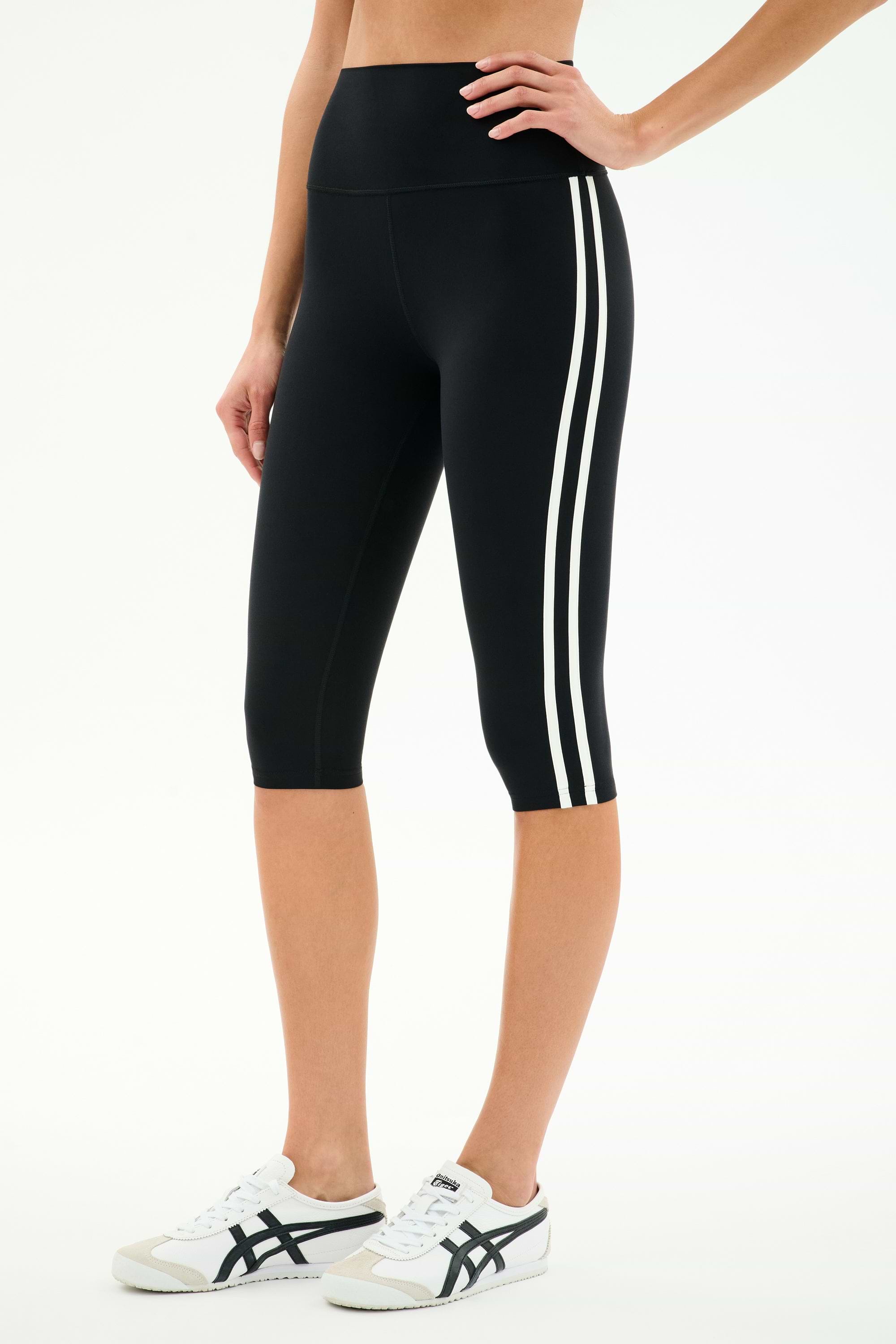 A person wears SPLITS59 Ella High Waist Airweight Capri in Black/White, featuring two white side stripes, paired with white sneakers with black accents, standing against a plain white background.