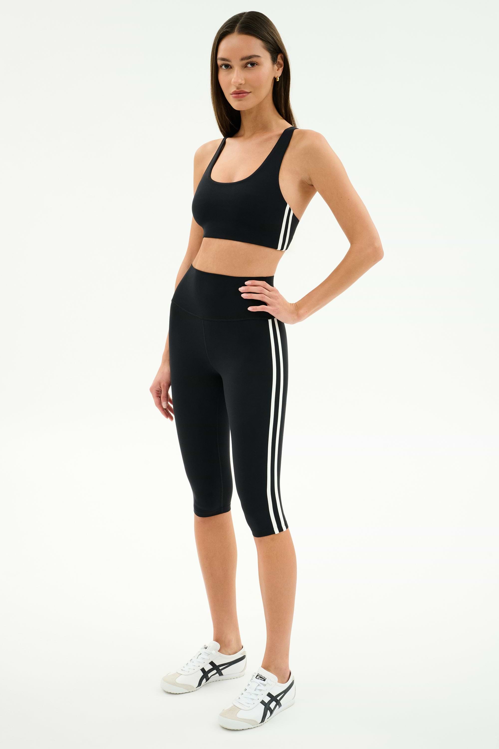A woman stands in a studio wearing the SPLITS59 Ella High Waist Airweight Capri in Black/White, paired with a black sports bra and white athletic sneakers.