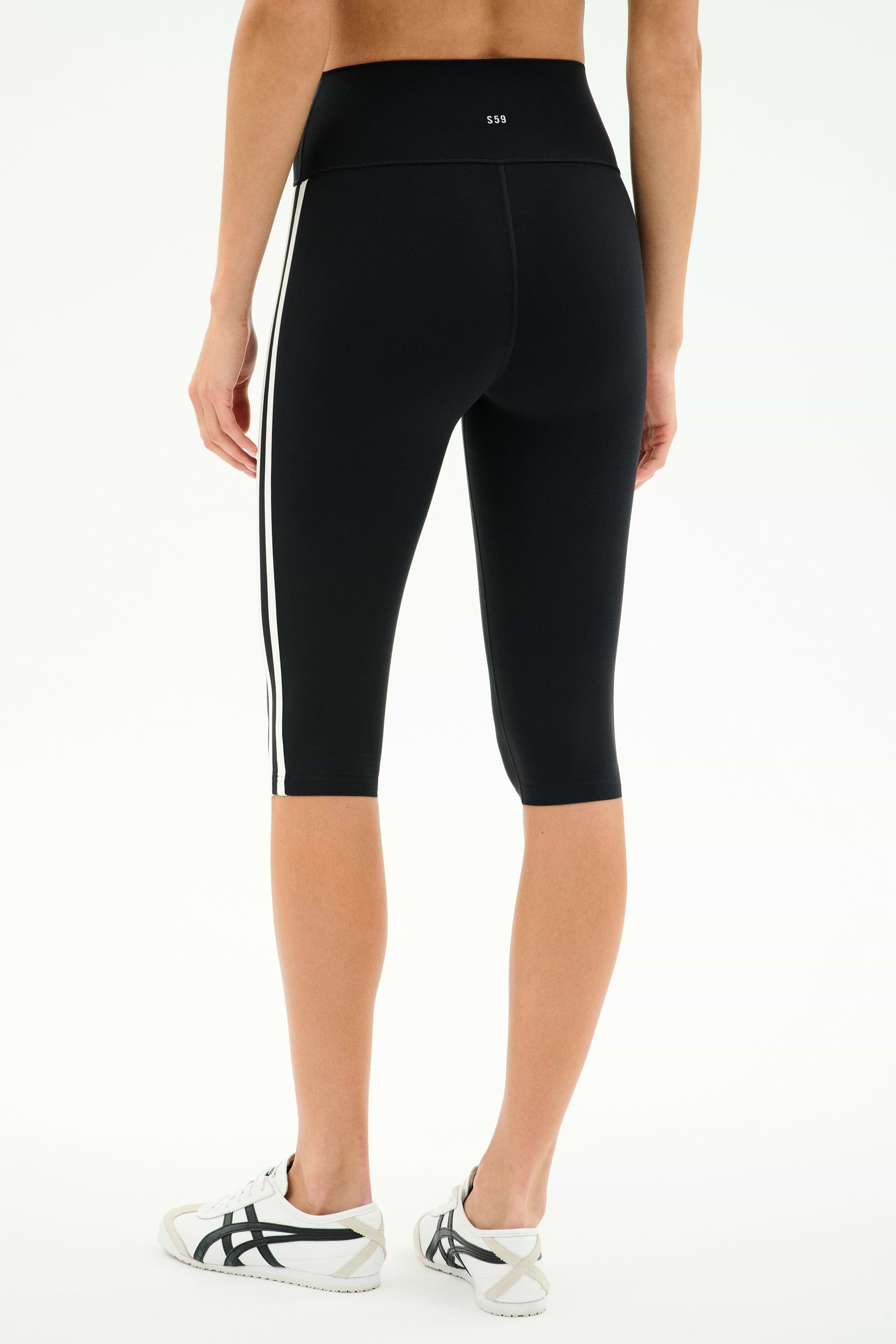 Model wears SPLITS59 Ella High Waist Airweight Capri in Black/White, featuring white side stripes, paired with white sneakers. Shown from the back against a plain background—sleek style and comfort for any workout.
