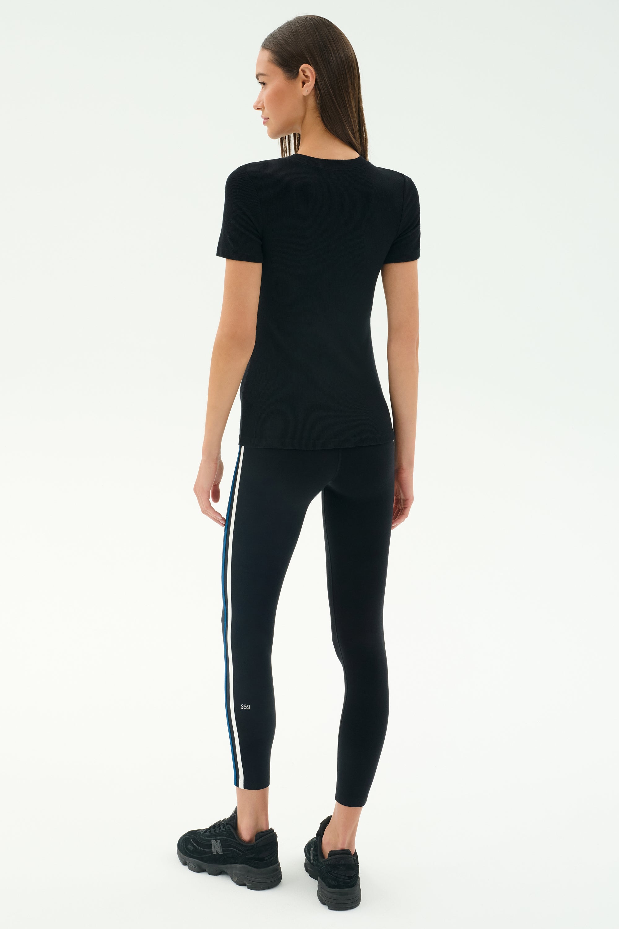 A woman faces away wearing the SPLITS59 Louise Rib Short Sleeve in black, paired with black leggings featuring white and blue side stripes, and black sneakers against a plain light background.
