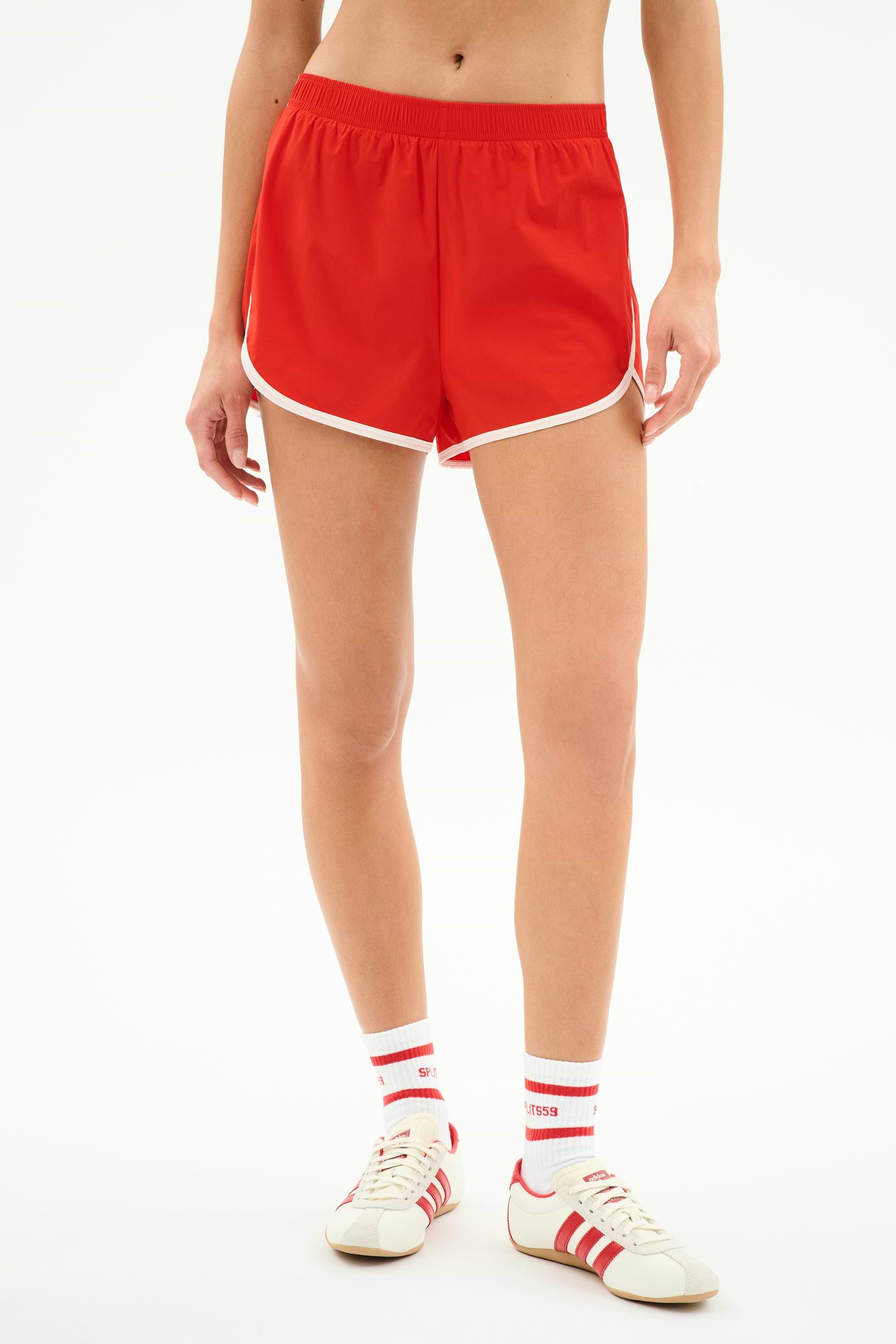 Model wears SPLITS59 Megan Woven Nylon Run Short in Lotus Red/Creme with white socks featuring red stripes and white sneakers with red accents, standing against a plain white background.