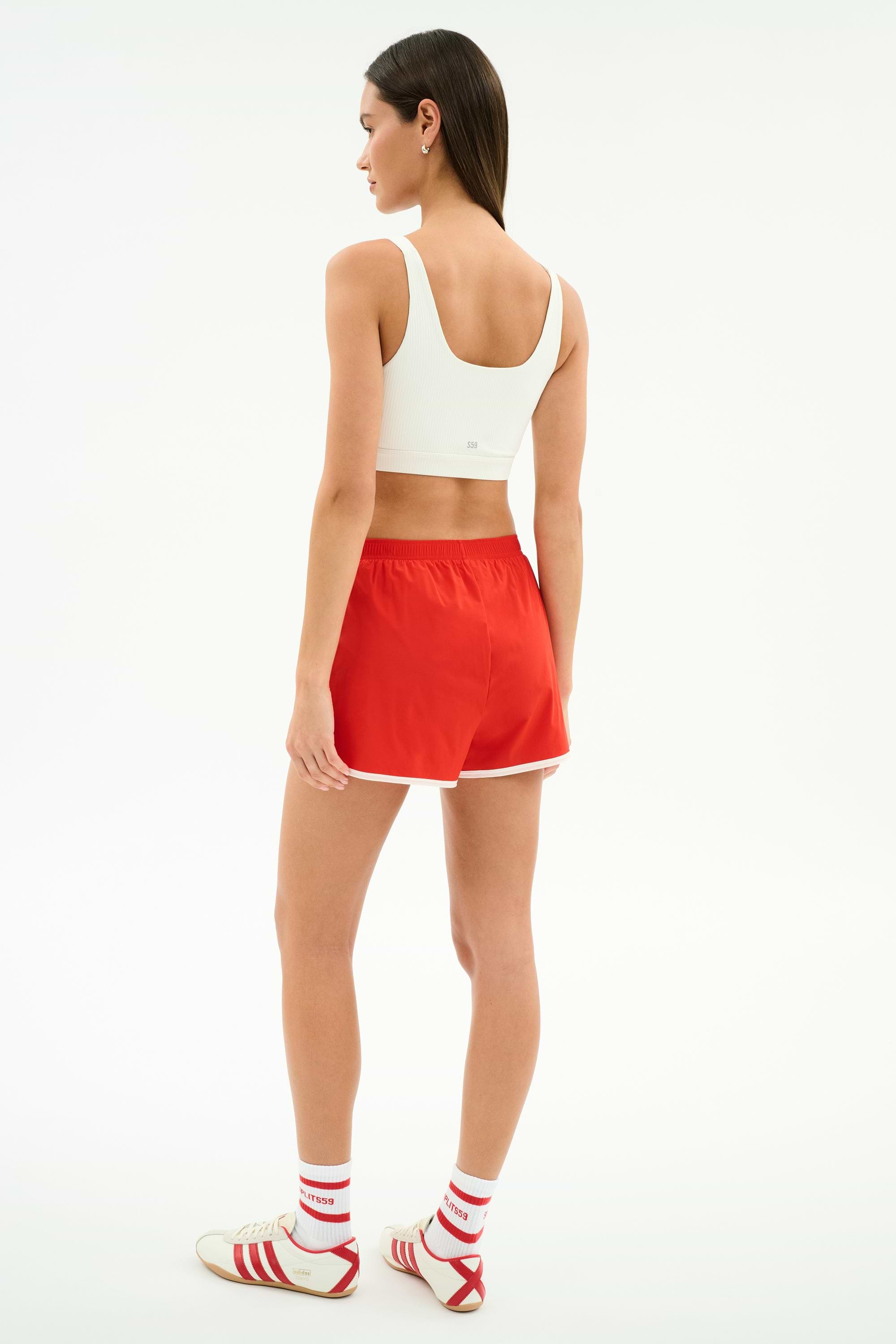 A woman faces away, wearing a white spandex sports bra and SPLITS59 Megan Woven Nylon Run Short in Lotus Red/Creme, paired with white socks with red stripes and white sneakers with red accents.