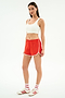 A woman wears a white spandex crop top, SPLITS59 Megan Woven Nylon Run Short in Lotus Red/Creme, white socks, and white sneakers with red stripes against a plain white background.