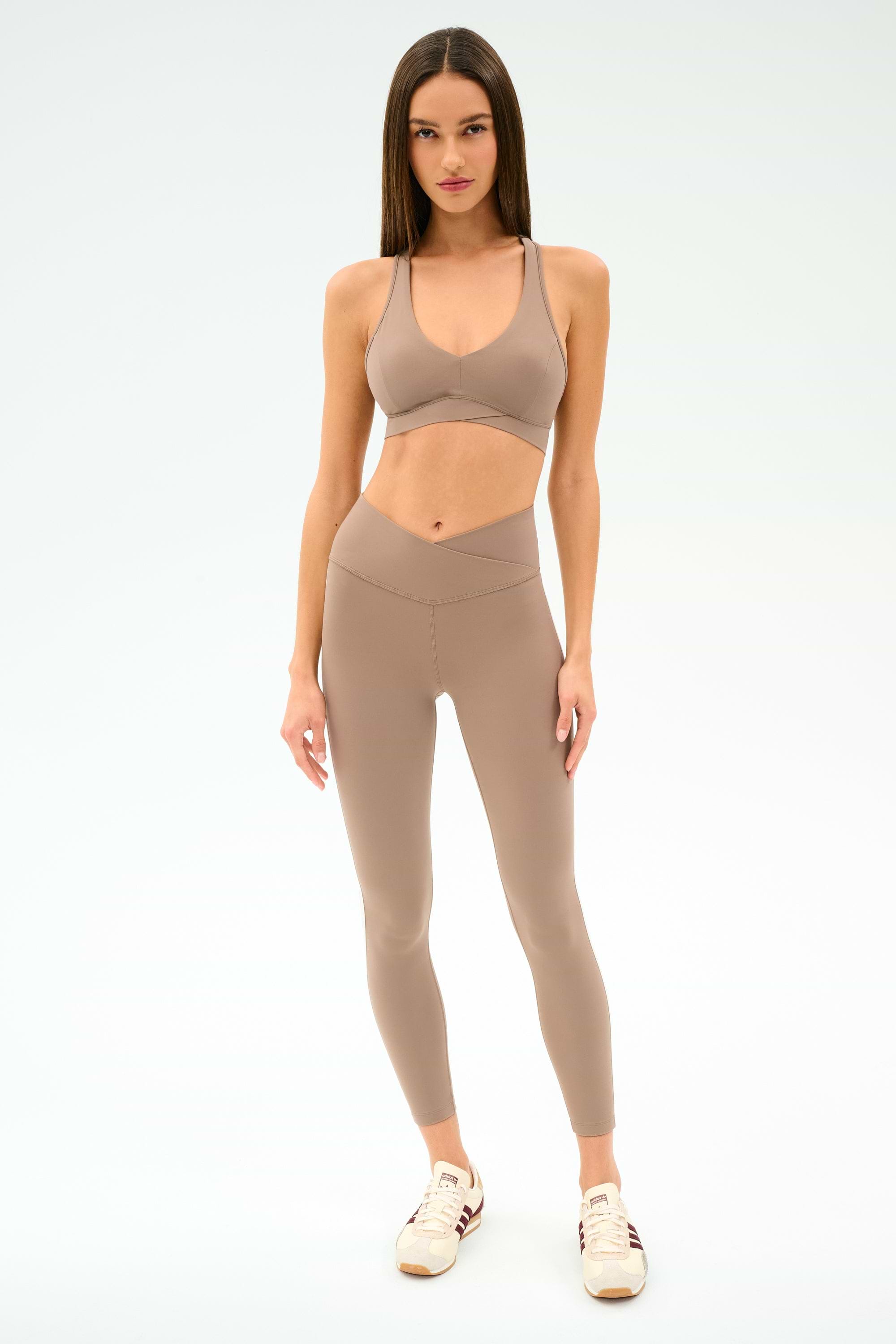 A woman stands against a plain background wearing the SPLITS59 Mia Rigor Legging w/ Stripe in Latte/White, paired with a taupe sports bra and beige sneakers.