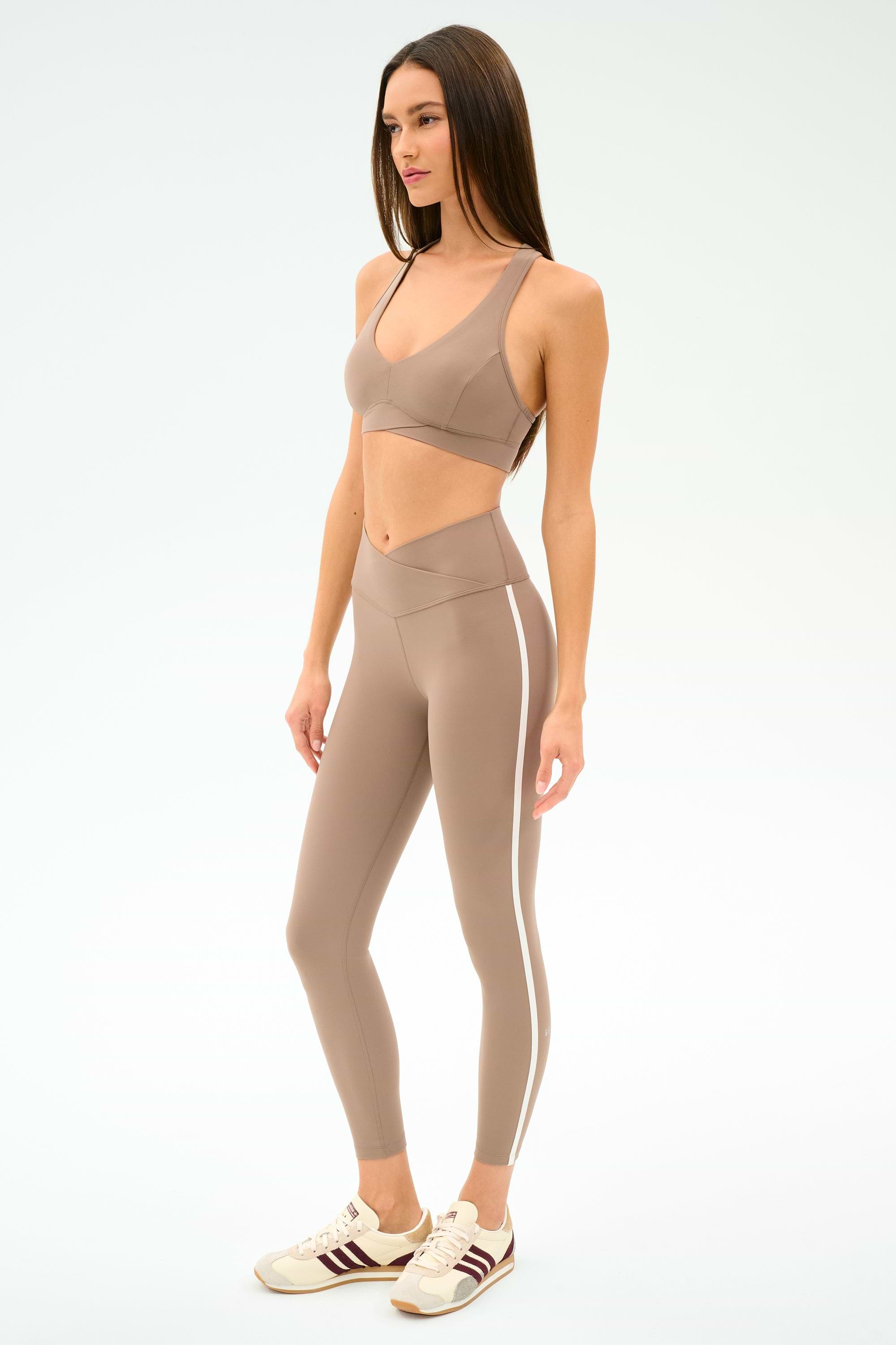 A woman poses against a plain light background wearing the SPLITS59 Mia Rigor Bra in Latte and high-waisted leggings with white side stripes, paired with cream and maroon sneakers.