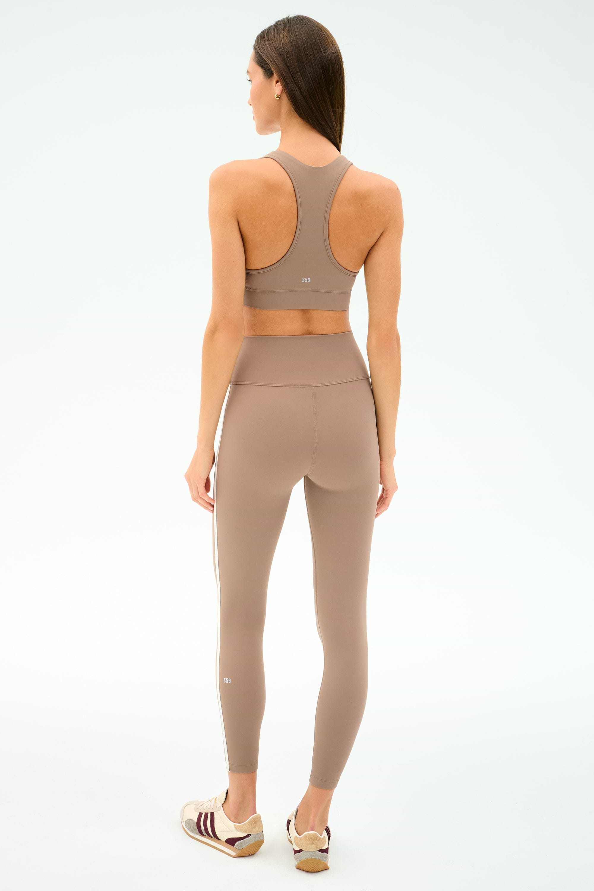 A woman faces away wearing SPLITS59’s Mia Rigor Legging w/ Stripe in Latte/White with a matching sports bra and white-maroon sneakers, set against a plain white background.