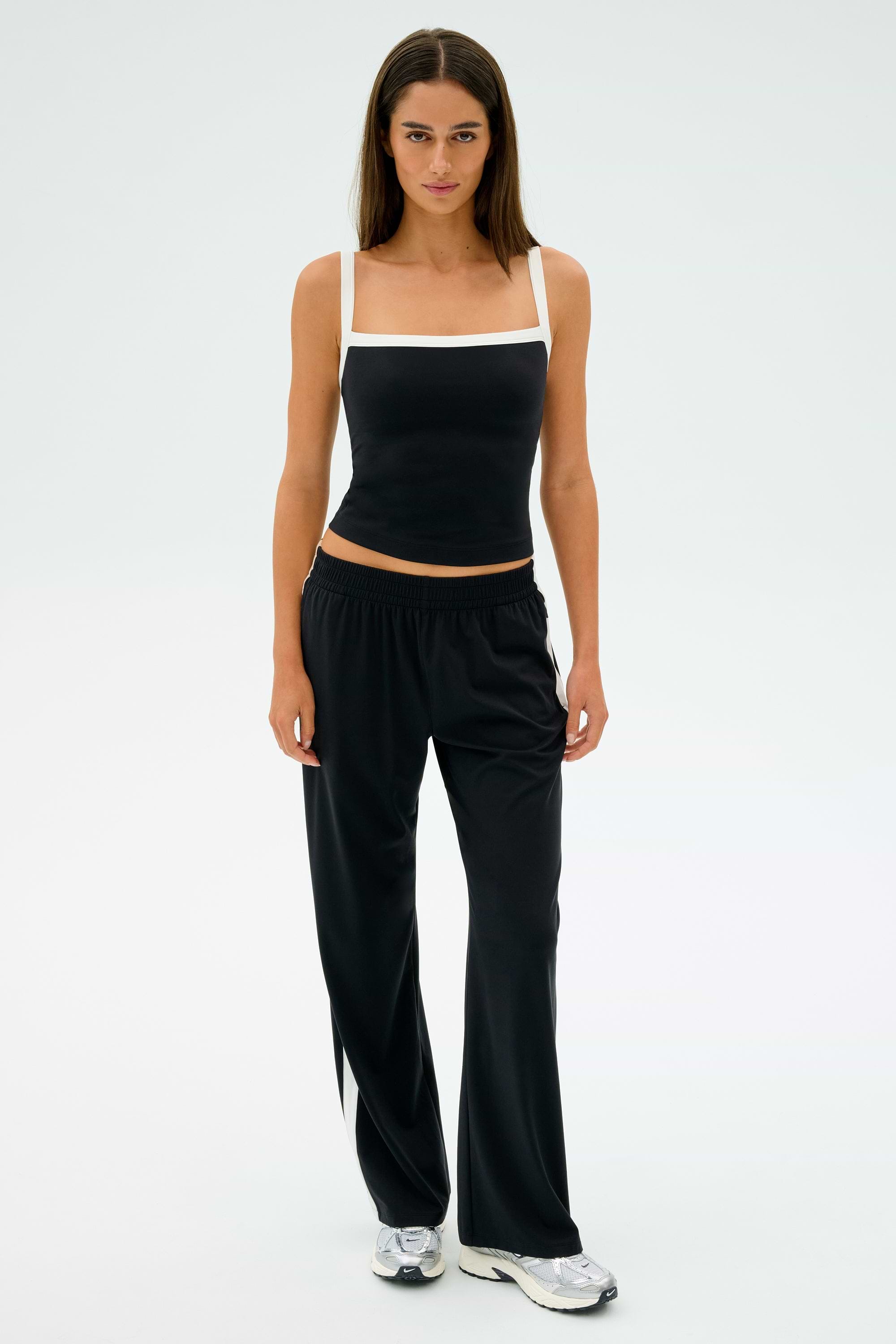 A person is standing against a plain background, wearing a black tank top, SPLITS59's Maxie Rigor Track Pant in Black/White, and silver sneakers.