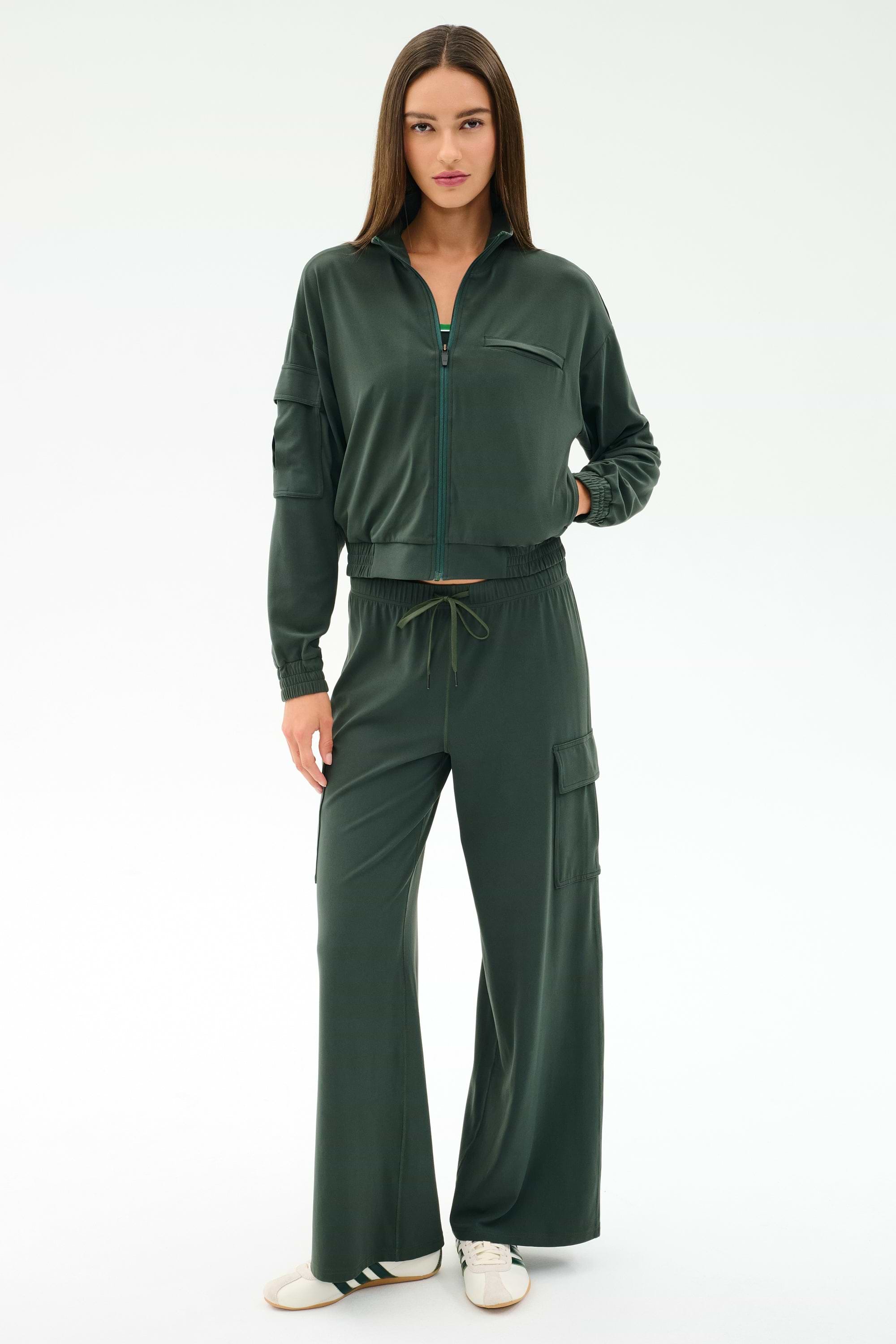 A woman faces forward in SPLITS59’s Alex Airweight Cargo Pant - Military, styled with a dark green nylon zip-up jacket and white sneakers, against a plain white background.