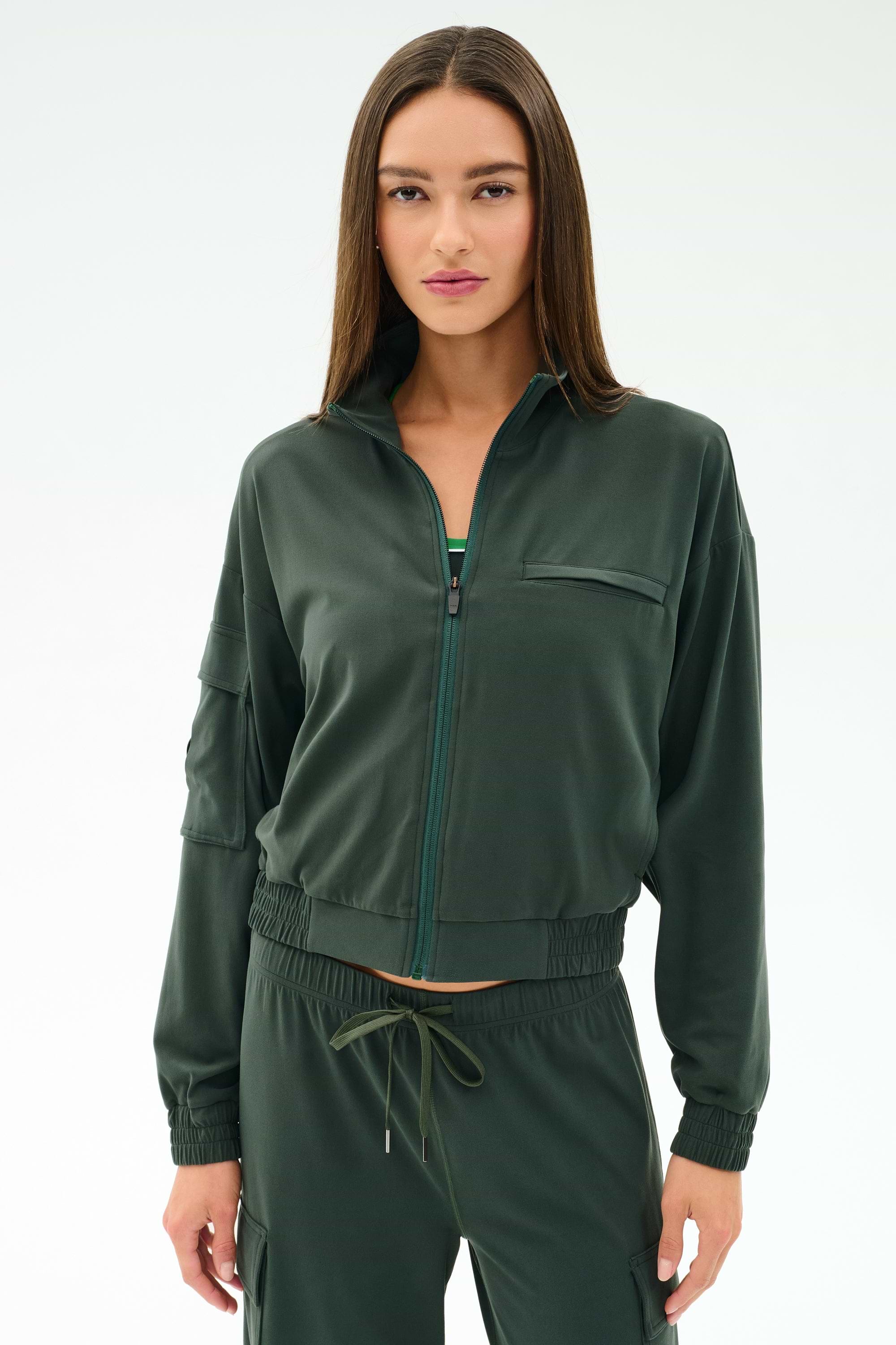 A woman wears the SPLITS59 Peaches Airweight Cargo Jacket in Military, styled with matching drawstring pants, both made from soft nylon spandex, standing against a plain light background.
