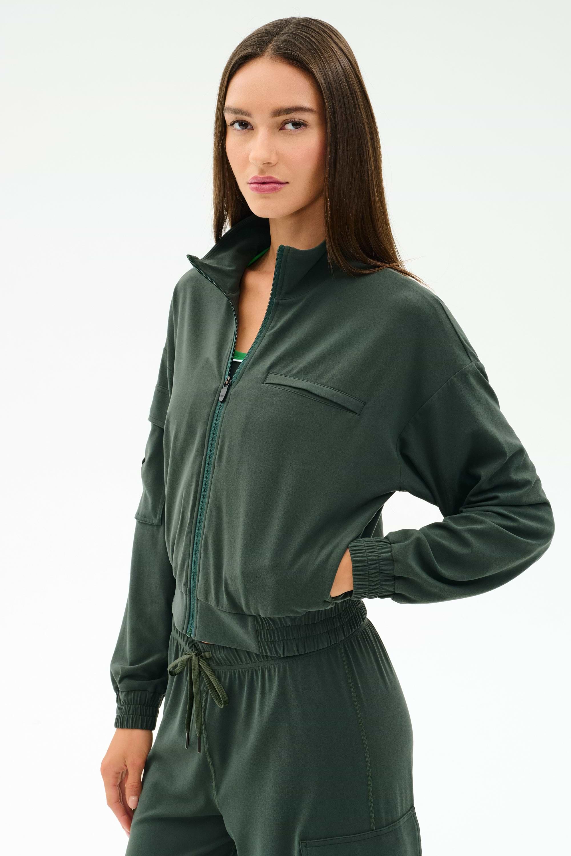 A woman wears the SPLITS59 Peaches Airweight Cargo Jacket in Military, made from soft nylon spandex, with her hand in the pocket, standing against a plain light background.