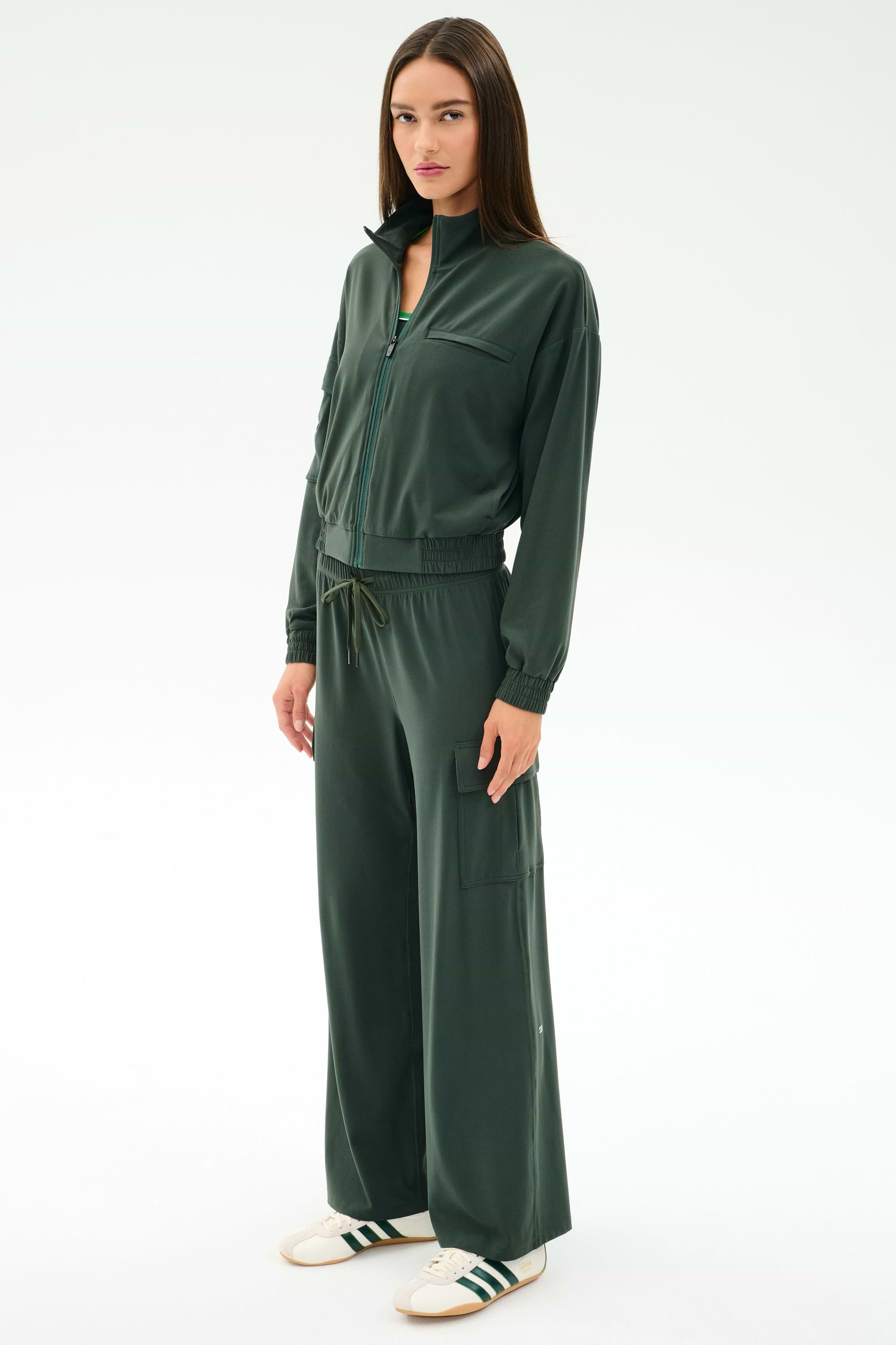 A woman faces forward wearing SPLITS59's Alex Airweight Cargo Pant in Military, paired with a dark green nylon zip-up jacket and white sneakers, standing against a plain white background.