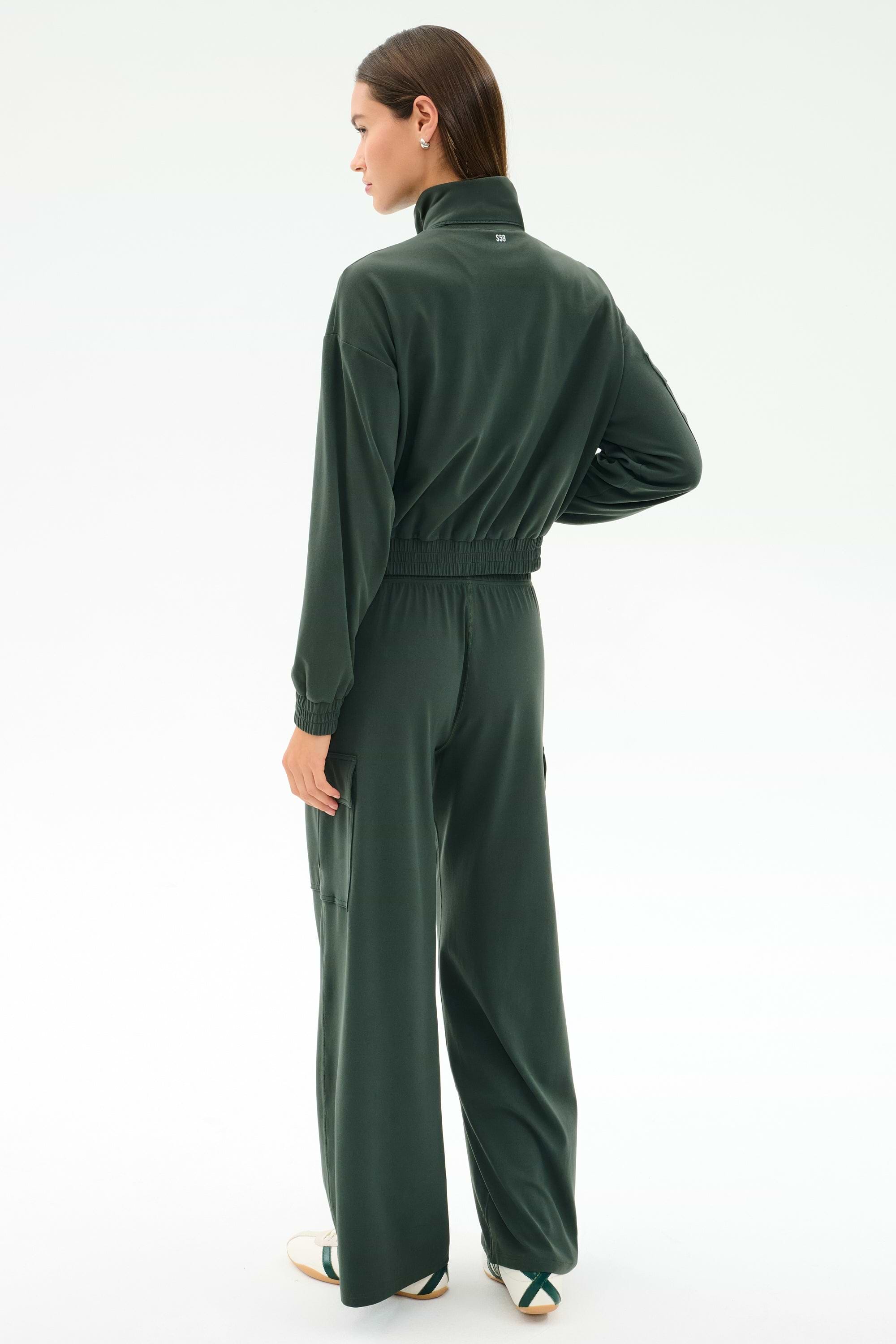 A woman stands with her back to the camera wearing SPLITS59's Alex Airweight Cargo Pant in Military, styled with a dark green top and white sneakers, on a plain white background.