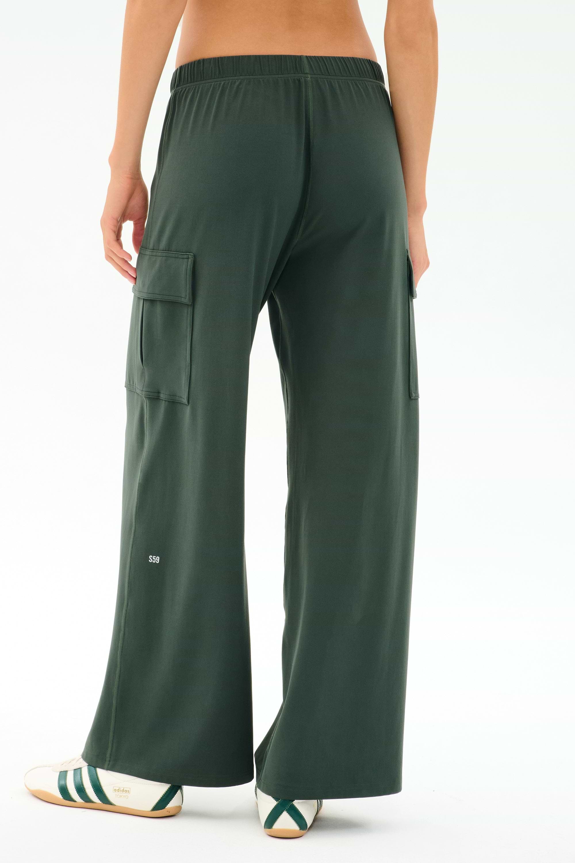Model wearing SPLITS59’s Alex Airweight Cargo Pant in Military, featuring wide legs and side pockets, paired with white slip-on shoes with green stripes; shown from the back against a plain background.