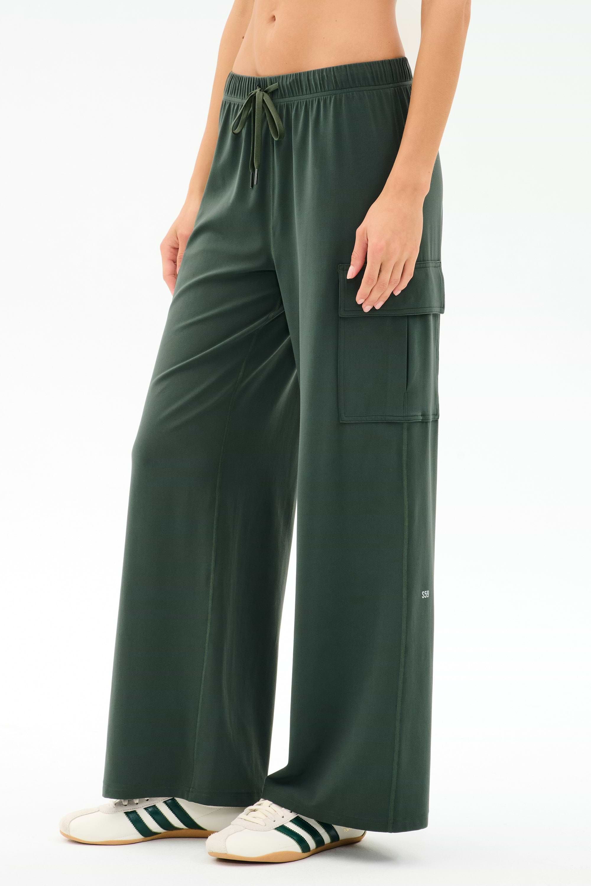 A person wears SPLITS59’s Alex Airweight Cargo Pant in Military, featuring a soft nylon blend, paired with white sneakers with green stripes, standing against a plain light background.